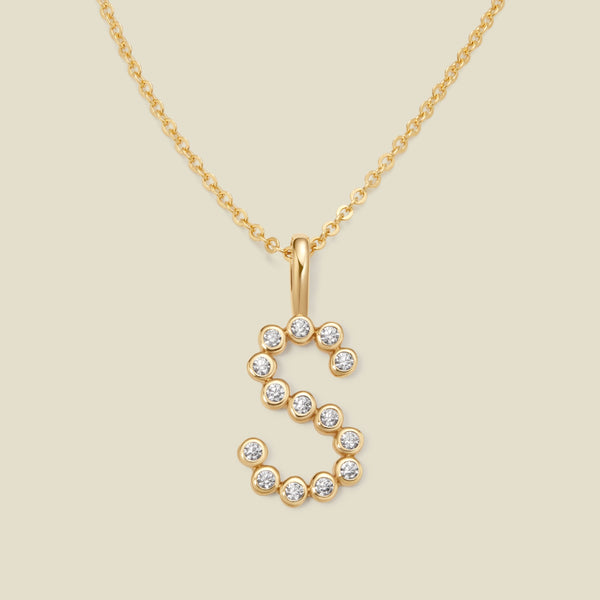 A gold pendant chain necklace with a gold uppercase letter 'S' charm made up of round bezel-set clear gemstones, set against a solid beige background.