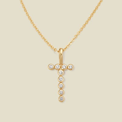 A gold pendant chain necklace with a gold uppercase letter 'T' charm made up of round bezel-set clear gemstones, set against a solid beige background.