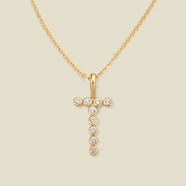 A gold pendant chain necklace with a gold uppercase letter 'T' charm made up of round bezel-set clear gemstones, set against a solid beige background.