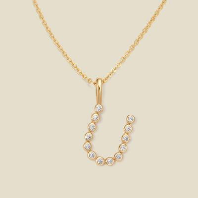 A gold pendant chain necklace with a gold uppercase letter 'U' charm made up of round bezel-set clear gemstones, set against a solid beige background.