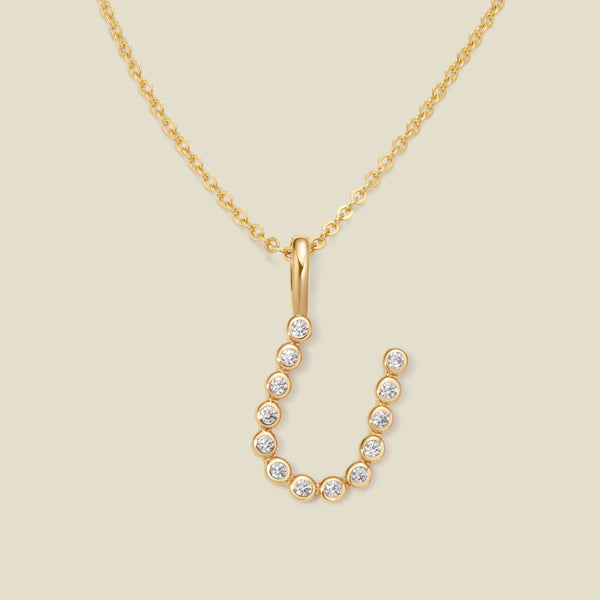 A gold pendant chain necklace with a gold uppercase letter 'U' charm made up of round bezel-set clear gemstones, set against a solid beige background.