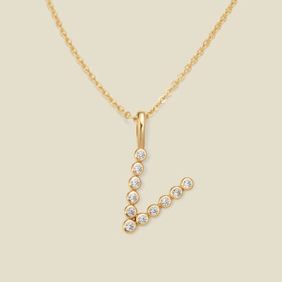 A gold pendant chain necklace with a gold uppercase letter 'V' charm made up of round bezel-set clear gemstones, set against a solid beige background.
