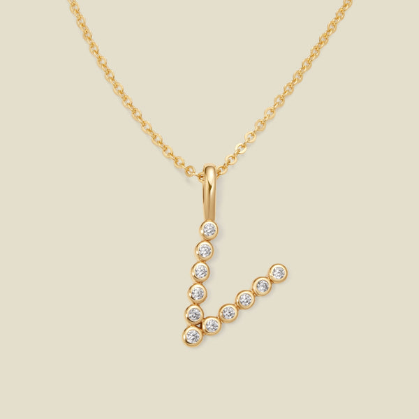 A gold pendant chain necklace with a gold uppercase letter 'V' charm made up of round bezel-set clear gemstones, set against a solid beige background.
