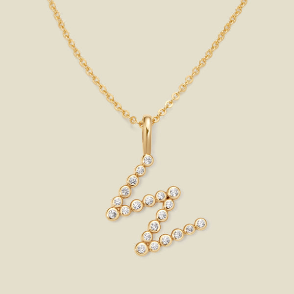 A gold pendant chain necklace with a gold uppercase letter 'W' charm made up of round bezel-set clear gemstones, set against a solid beige background.