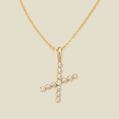 A gold pendant chain necklace with a gold uppercase letter 'X' charm made up of round bezel-set clear gemstones, set against a solid beige background.