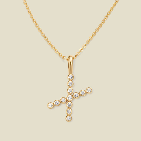 A gold pendant chain necklace with a gold uppercase letter 'X' charm made up of round bezel-set clear gemstones, set against a solid beige background.