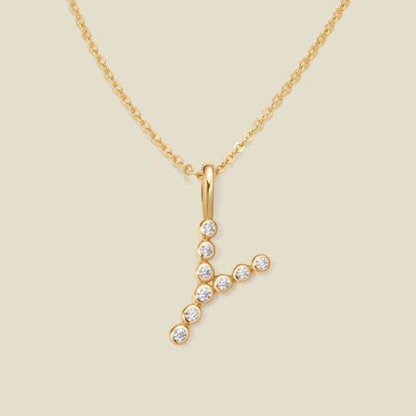 A gold pendant chain necklace with a gold uppercase letter 'Y' charm made up of round bezel-set clear gemstones, set against a solid beige background.