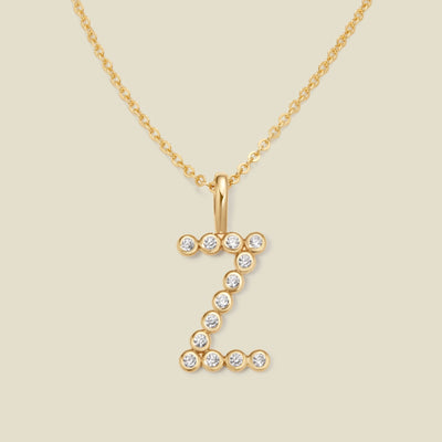 A gold pendant chain necklace with a gold uppercase letter 'Z' charm made up of round bezel-set clear gemstones, set against a solid beige background.