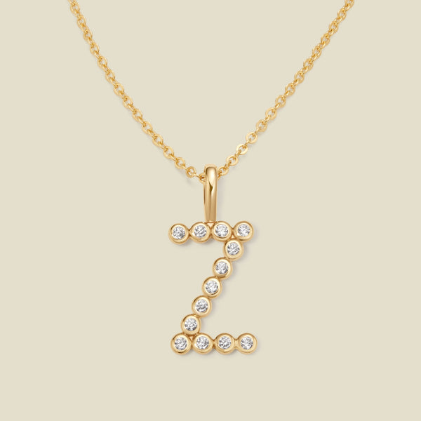 A gold pendant chain necklace with a gold uppercase letter 'Z' charm made up of round bezel-set clear gemstones, set against a solid beige background.