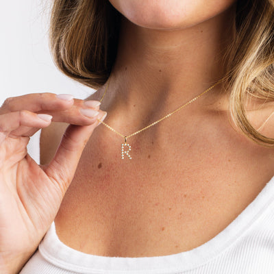 A close-up lifestyle shot of a person's chest with brunette hair, wearing a white round neck t-shirt holding a gold pendant necklace with the letter 'R'.