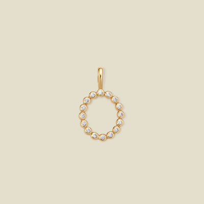 A gold initial 'O' charm featuring small clear round-cut stones, set against a solid light beige background.