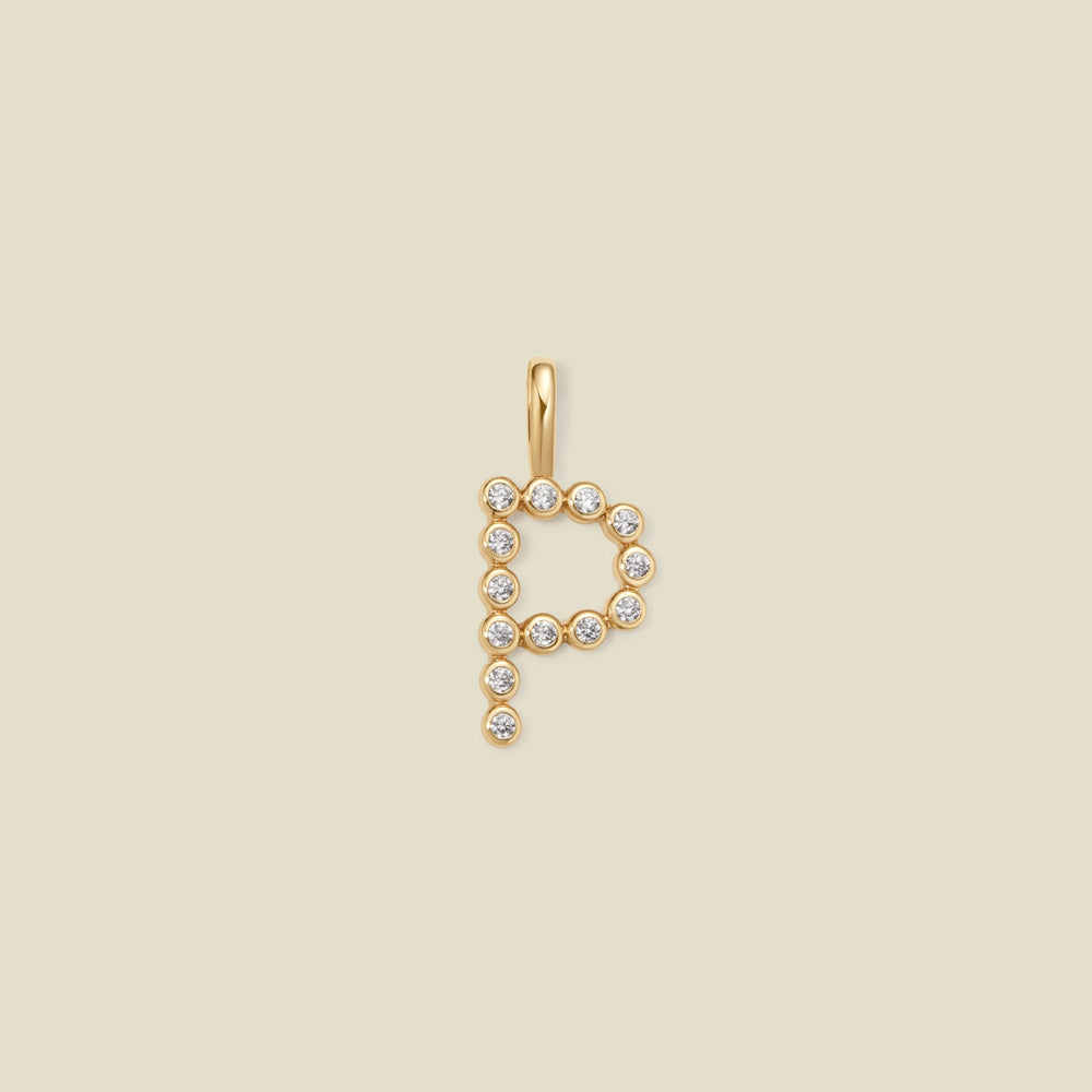 A gold letter 'P' pendant charm embellished with small clear round gemstones, centered on a solid light beige background.