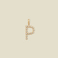 A gold letter 'P' pendant charm embellished with small clear round gemstones, centered on a solid light beige background.