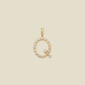 A gold letter 'Q' pendant charm embellished with small clear round gemstones, centered on a solid light beige background.