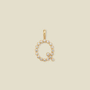 A gold letter 'Q' pendant charm embellished with small clear round gemstones, centered on a solid light beige background.