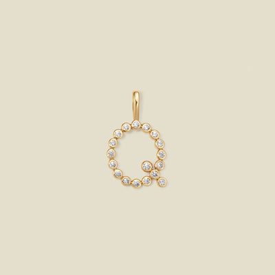 A gold letter 'Q' pendant charm embellished with small clear round gemstones, centered on a solid light beige background.
