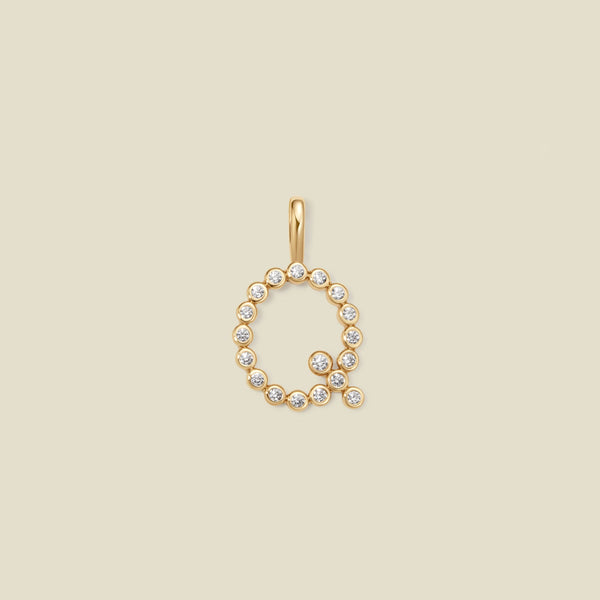 A gold letter 'Q' pendant charm embellished with small clear round gemstones, centered on a solid light beige background.