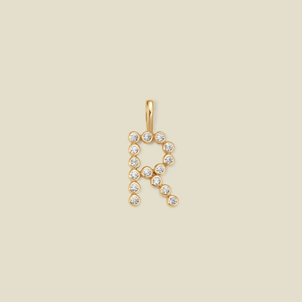 A gold letter 'R' pendant charm embellished with small clear round gemstones, centered on a solid light beige background.