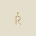 A gold letter 'R' pendant charm embellished with small clear round gemstones, centered on a solid light beige background.