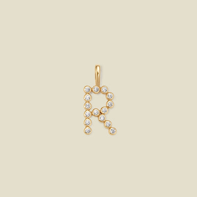 A gold letter 'R' pendant charm embellished with small clear round gemstones, centered on a solid light beige background.