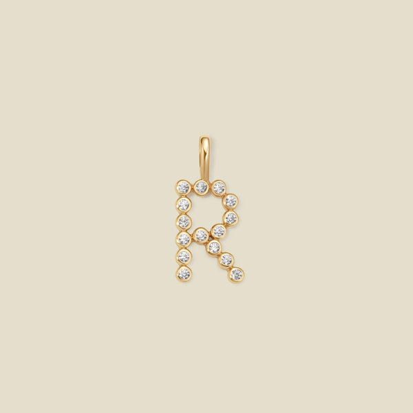 A gold letter 'R' pendant charm embellished with small clear round gemstones, centered on a solid light beige background.