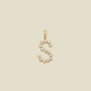 A gold letter 'S' pendant charm embellished with small clear round gemstones, centered on a solid light beige background.