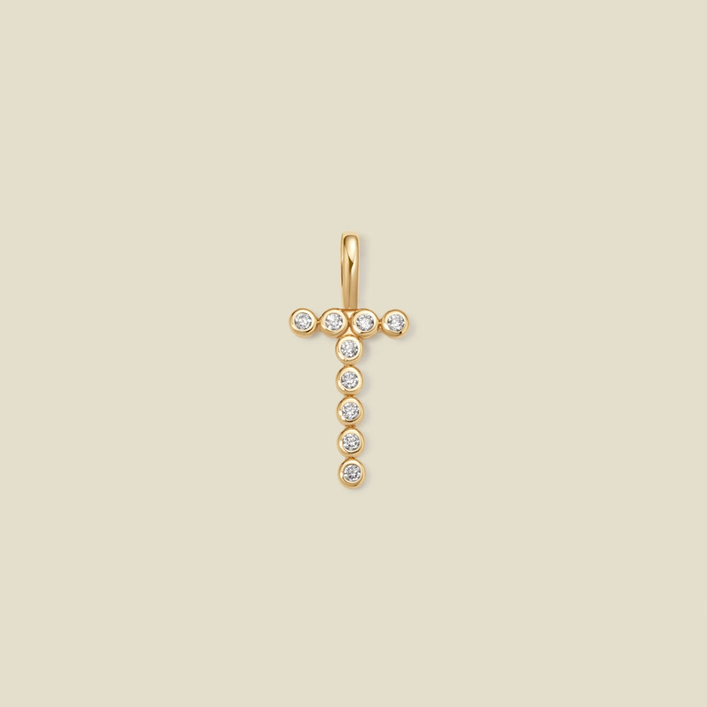 A gold initial 'T' charm comprised of clear round bezel-set gemstones, set against a solid beige background.