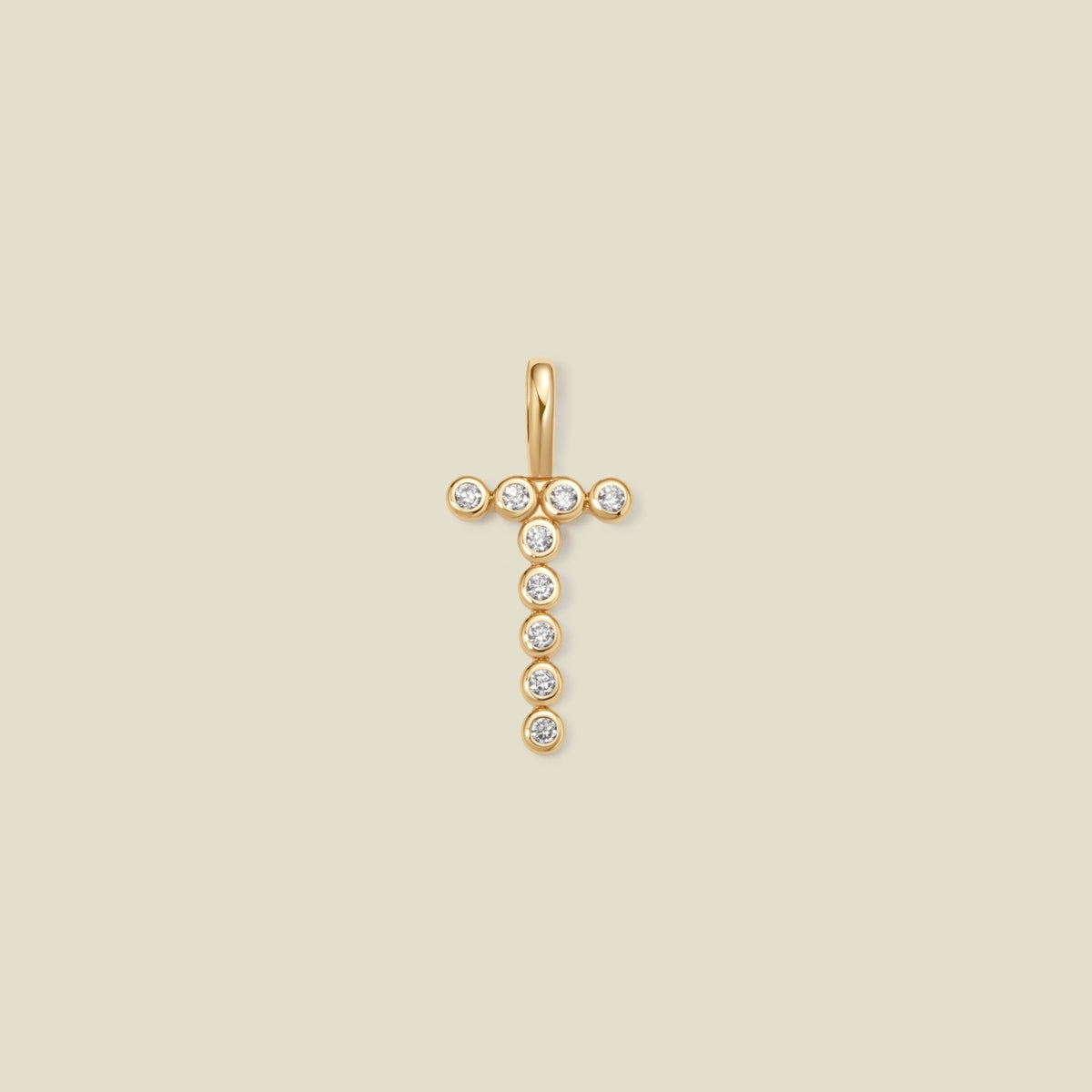 A gold initial 'T' charm comprised of clear round bezel-set gemstones, set against a solid beige background.