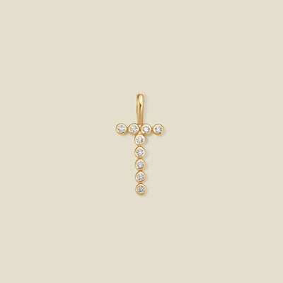 A gold initial 'T' charm comprised of clear round bezel-set gemstones, set against a solid beige background.