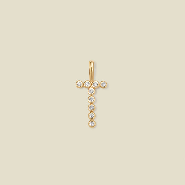 A gold initial 'T' charm comprised of clear round bezel-set gemstones, set against a solid beige background.