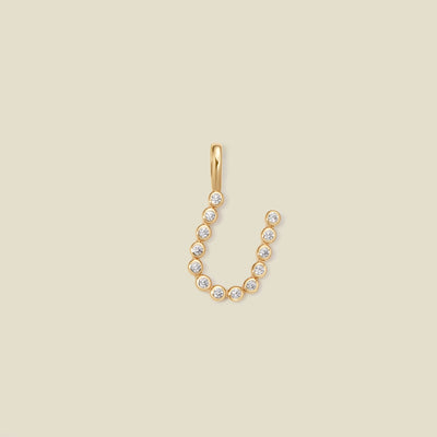 A gold initial 'U' charm made up of small round bezel-set clear stones, set against a solid beige background.