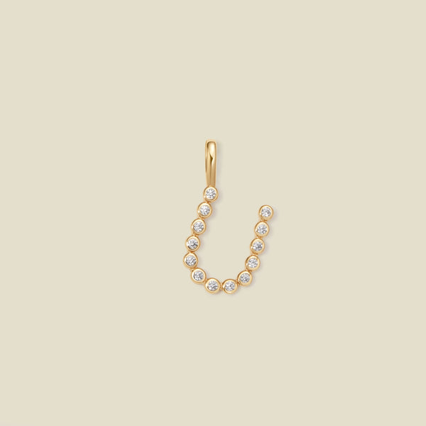 A gold initial 'U' charm made up of small round bezel-set clear stones, set against a solid beige background.