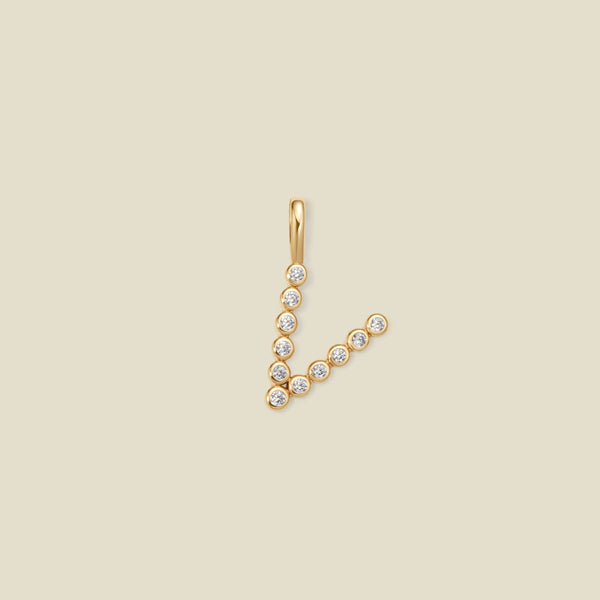 A gold initial pendant charm shaped like the letter 'V', adorned with small clear round gemstones, shown on a solid light beige background.