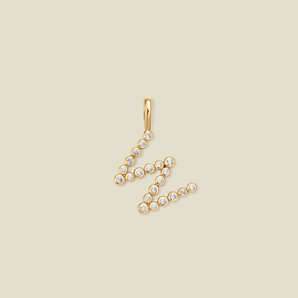 A gold letter 'W' pendant charm embellished with small clear round gemstones, set against a solid light beige background.