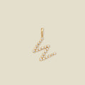 A gold letter 'W' pendant charm embellished with small clear round gemstones, set against a solid light beige background.