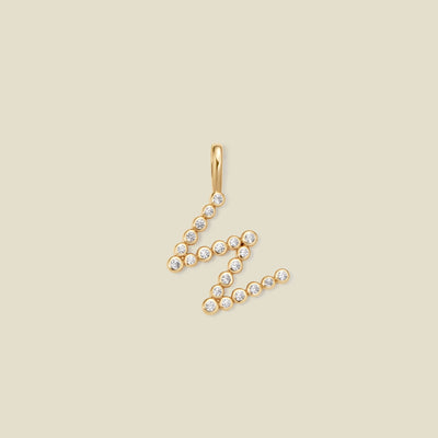 A gold letter 'W' pendant charm embellished with small clear round gemstones, set against a solid light beige background.