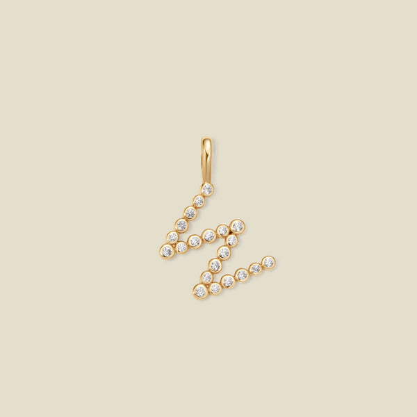 A gold letter 'W' pendant charm embellished with small clear round gemstones, set against a solid light beige background.