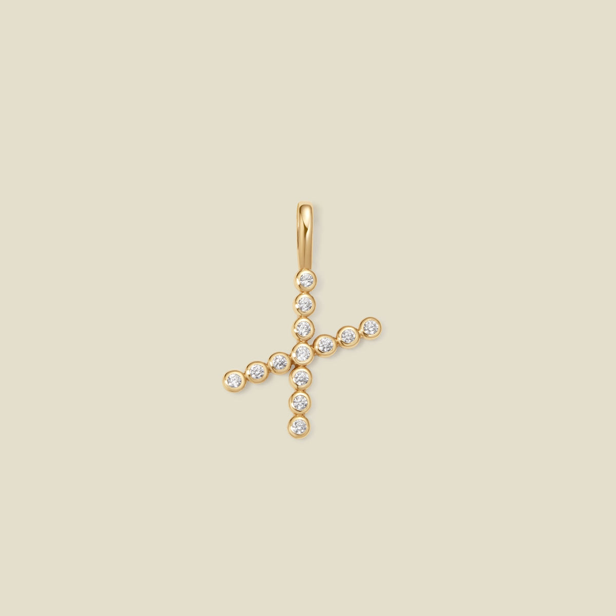A gold initial 'X' charm made up of small round bezel-set clear gemstones, set against a solid beige background.