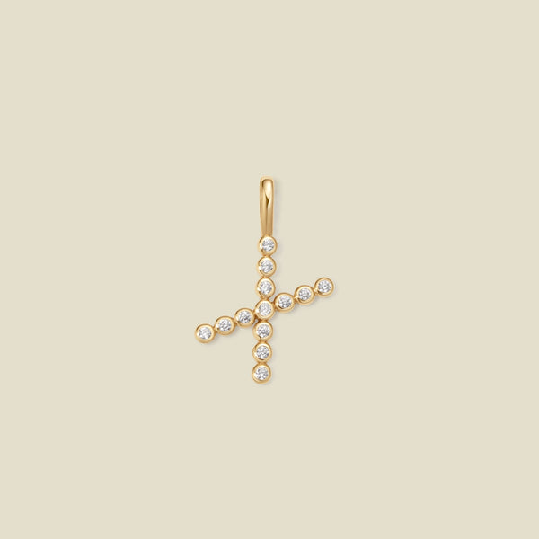 A gold initial 'X' charm made up of small round bezel-set clear gemstones, set against a solid beige background.