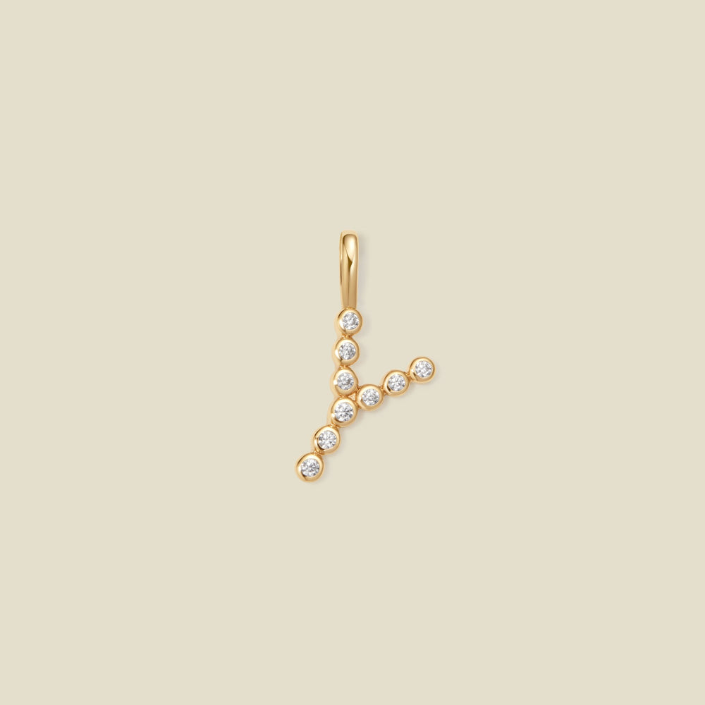 A gold initial pendant 'Y' charm, adorned with small clear round-cut gemstones, set against a solid light beige background.