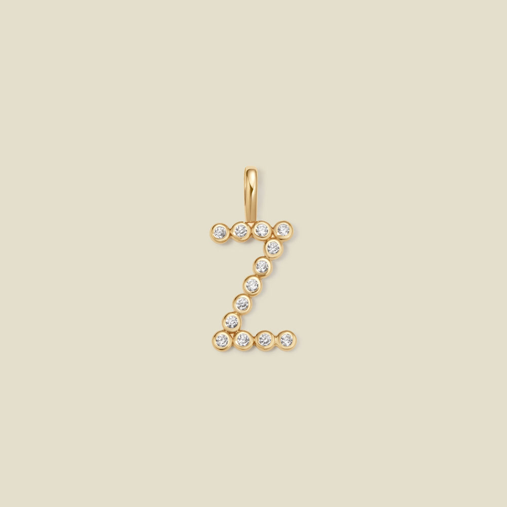 A gold initial 'Z' charm made up of small round bezel-set clear gemstones, set against a solid beige background.