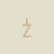 A gold initial 'Z' charm made up of small round bezel-set clear gemstones, set against a solid beige background.