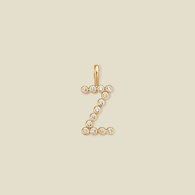 A gold initial 'Z' charm made up of small round bezel-set clear gemstones, set against a solid beige background.