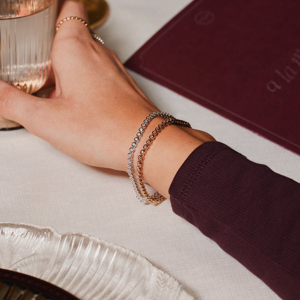 A close-up of a persons wrist resting on a restaurant table with a menu in the background, holding a glass and showcasing a gold and silver tennis bracelet with round bezel-set clear gemstones. 