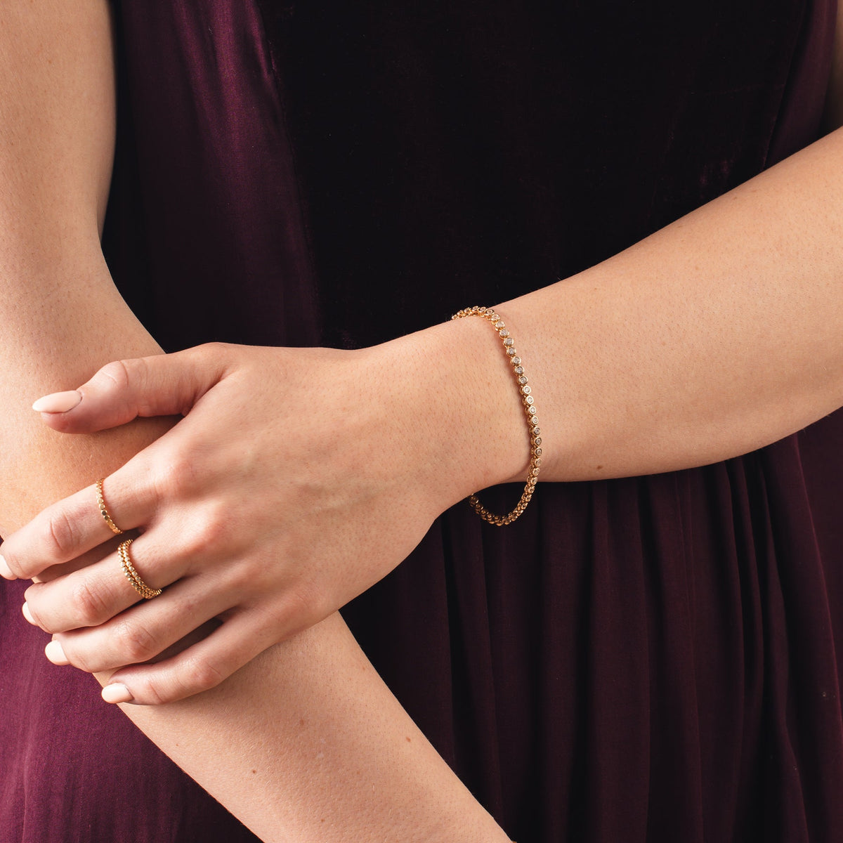 A close-up of a person's wrist wearing a gold tennis bracelet with round clear bezel-set gemstones and three matching gold rings, paired with a maroon dress.