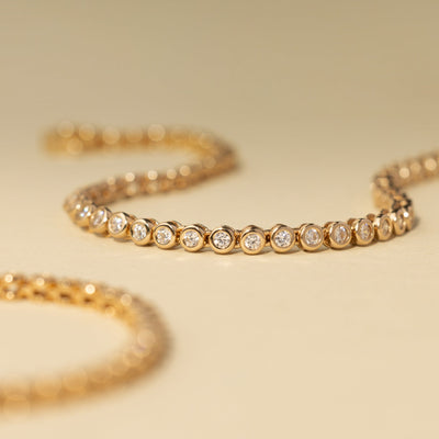 A gold tennis bracelet with bezel-set round clear gemstones, set on a solid beige background with another bracelet blurred in the foreground.