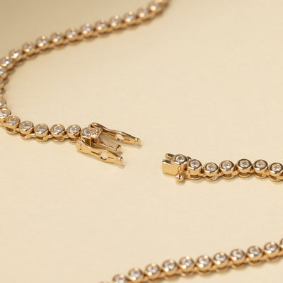 A box clasp laying open on a gold tennis bracelet with round bezel-set clear gemstones laying on a solid beige background.