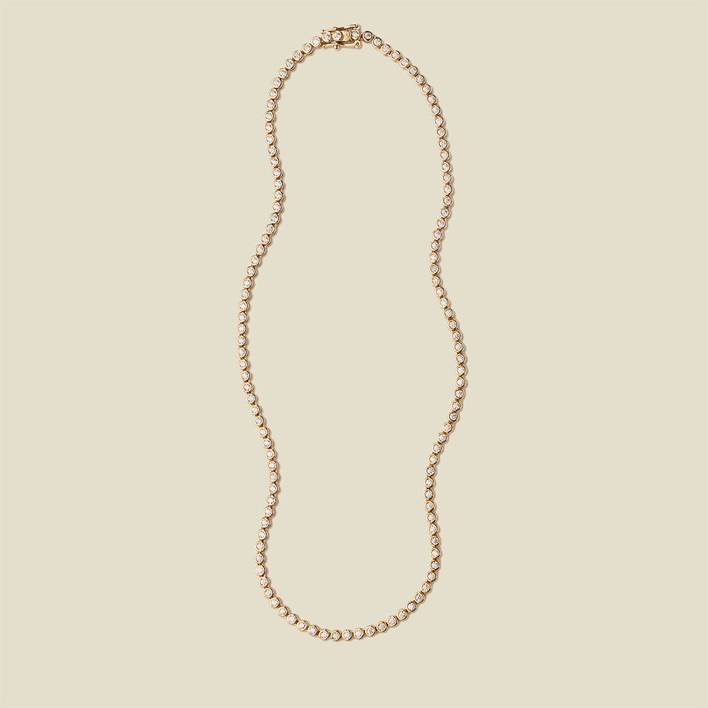 A gold tennis necklace with round bezel-set clear gemstones, set against a solid beige background.