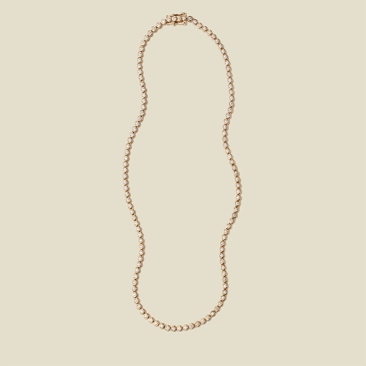 A gold tennis necklace with round bezel-set clear gemstones, set against a solid beige background.