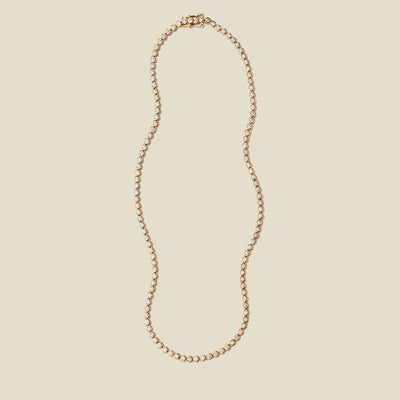 A gold tennis necklace with round bezel-set clear gemstones, set against a solid beige background.